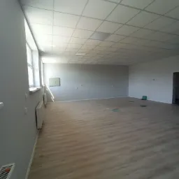 Wnętrze pustego, pomalowanego na szaro pomieszczenia w Bydgoszczy z panelami na podłodze i kasetonowym sufitem podwieszanym. Widoczne grzejniki przy ścianie z oknami i zielone elementy podłogowe.