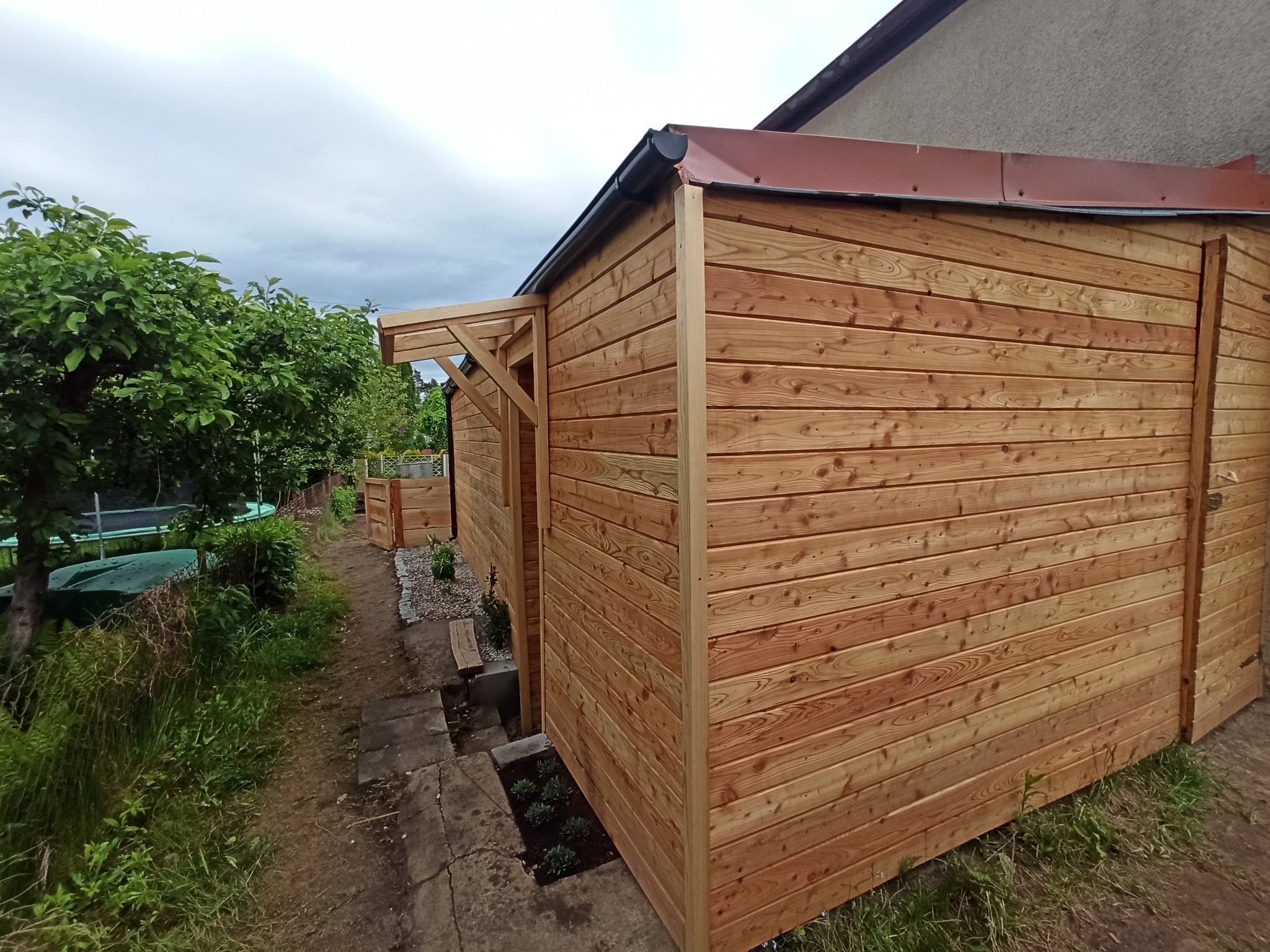 Nowa, drewniana konstrukcja z dachem, prawdopodobnie altana lub domek narzędziowy, z widocznymi elementami konstrukcyjnymi, przylegająca do budynku mieszkalnego, otoczona zielenią.