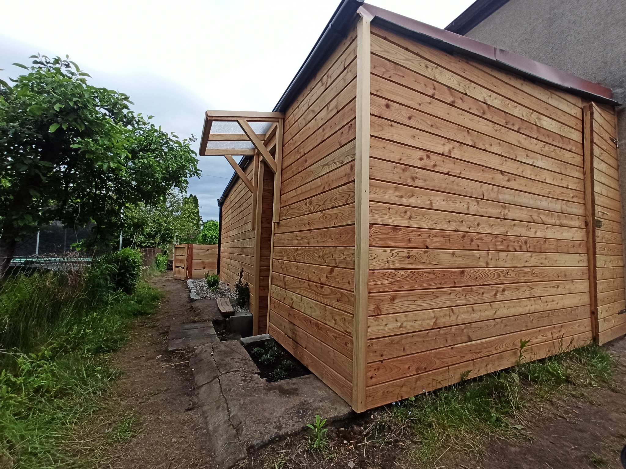 Nowoczesna zabudowa balkonu z jasnego drewna, zadaszenie nad wejściem, widok z zewnątrz na tle zieleni.