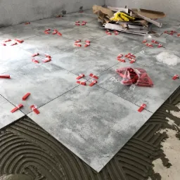 Układanie szarych płytek podłogowych z użyciem czerwonych klinów poziomujących, widoczna warstwa kleju i stos kartonów po płytkach w rogu pomieszczenia.