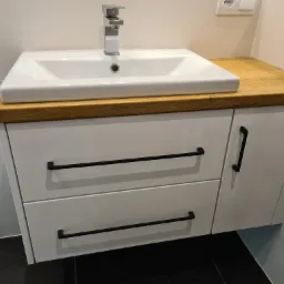 Biała szafka łazienkowa z dwiema szufladami i drewnianym blatem, na którym umieszczona jest biała umywalka z chromowaną baterią. Czarne, poziome uchwyty szuflad.
