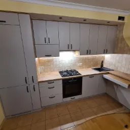 Jasnoszara zabudowa kuchenna z drewnianymi blatami, czarnymi uchwytami i wysuwanym blatem roboczym przy zlewozmywaku, nad płytą grzewczą wbudowane oświetlenie podszafkowe.