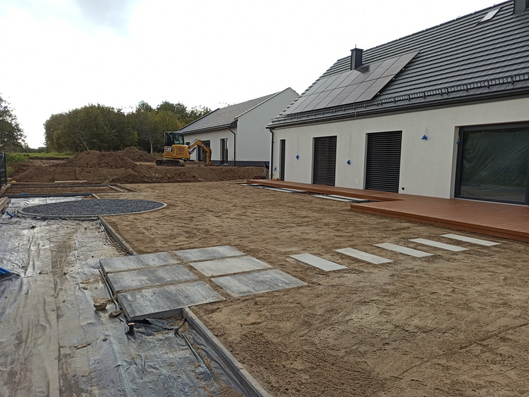 Niwelacja terenu pod ogród nowoczesnego domu z panelami słonecznymi, widoczny żółty mini-koparka CAT, przygotowane podłoże, ułożone płyty chodnikowe i obrzeża, wysypany kamień ozdobny.