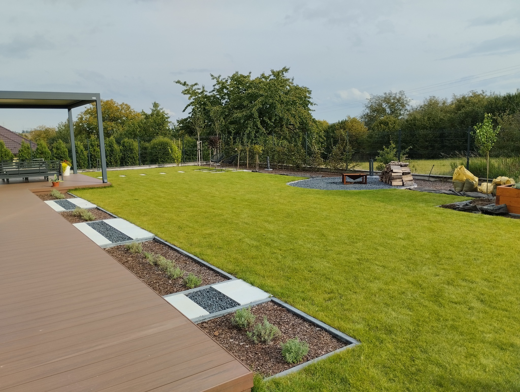 Widok na zadbany ogród z równo przystrzyżonym trawnikiem, drewnianym tarasem z pergolą, ścieżką z płyt i żwiru oraz elementami dekoracyjnymi, takimi jak ułożone drewno i młode drzewka.