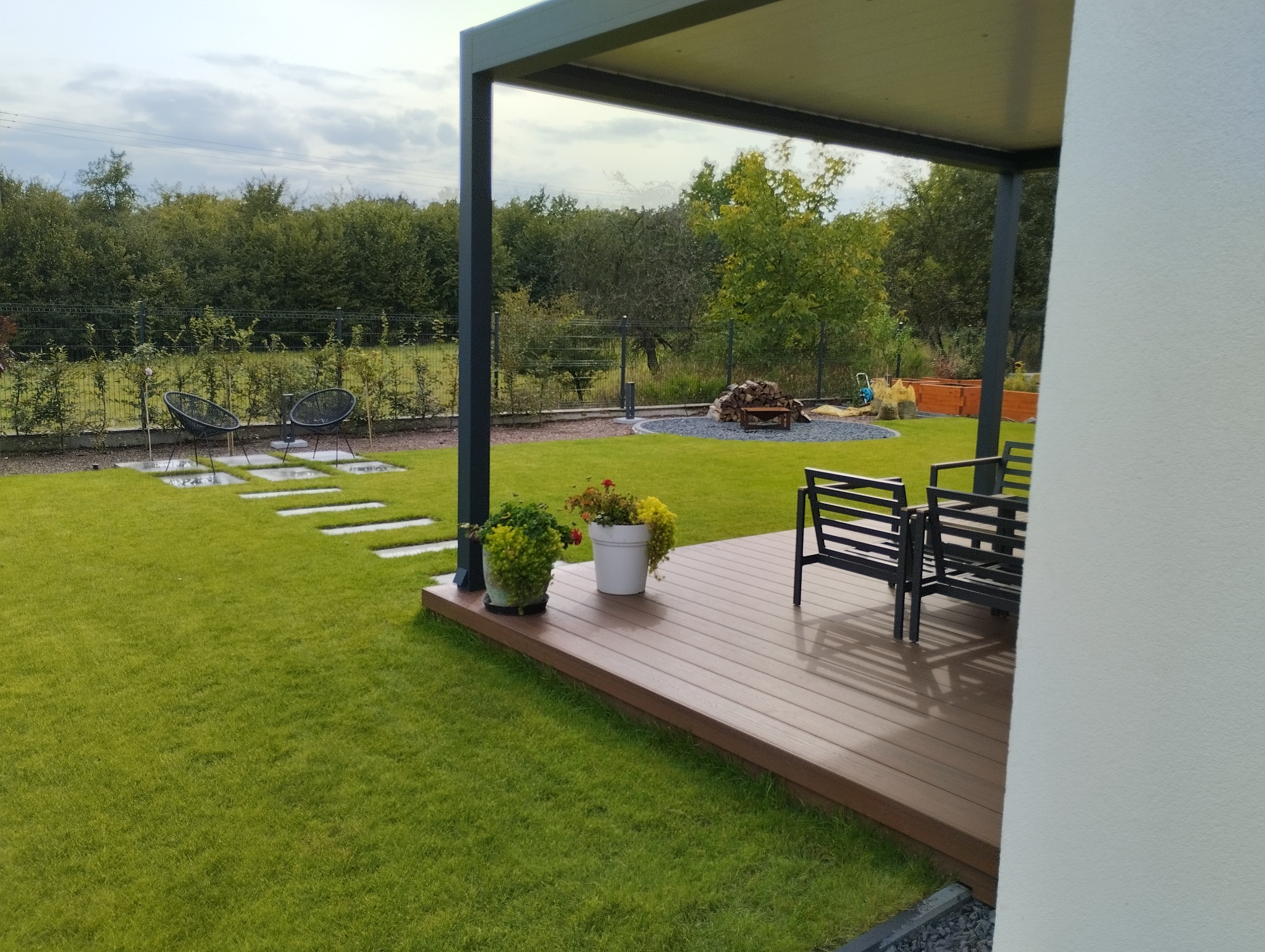 Nowoczesny ogród z równo przystrzyżonym trawnikiem, drewnianym tarasem z meblami ogrodowymi pod zadaszeniem, ścieżką z płyt betonowych i ozdobnym nasadzeniem z kamieni i drewna w tle.