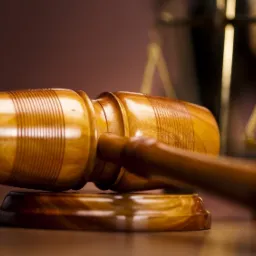 Drewniany młotek sędziowski spoczywający na podstawie, w tle rozmyte wagi sprawiedliwości, ciepłe oświetlenie.