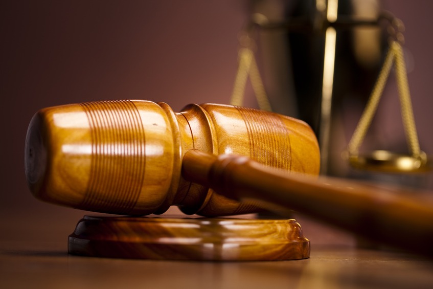 Drewniany młotek sędziowski spoczywający na podstawie, w tle rozmyte wagi sprawiedliwości, ciepłe oświetlenie.