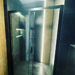 Nowoczesna kabina prysznicowa z przesuwnymi drzwiami w ciemnej ramie, podświetlona paskiem LED na ścianie z geometrycznym wzorem. W tle widoczne półki wnękowe.