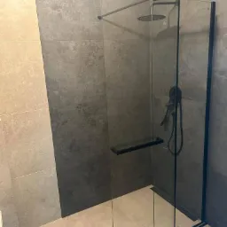 Nowoczesna kabina prysznicowa typu walk-in z czarnymi profilami i deszczownicą, szare płytki na ścianach o zróżnicowanej fakturze, jasna podłoga, minimalistyczny design.