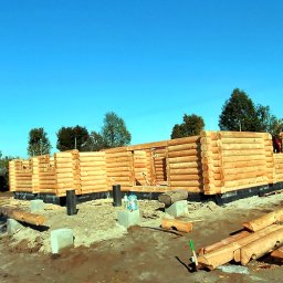 AFJ Domy z Bala Sp. z o.o. - Wznoszona konstrukcja z bali na fundamencie, widoczne elementy izolacji i przygotowane bale na pierwszym planie, błękitne niebo w tle.