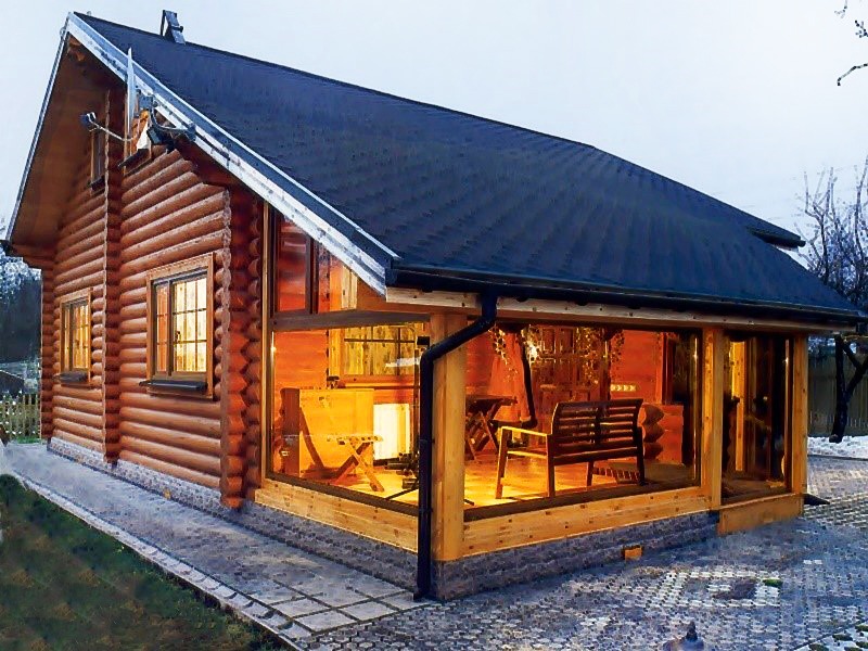 Zewnętrzny widok domu z bali z przeszklonym tarasem, oświetlonym ciepłym światłem, z widocznym fragmentem ogrodu i brukowanym chodnikiem.