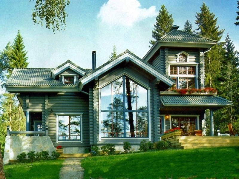 Dwukondygnacyjny dom z bali z dużymi oknami, zielonym dachem i drewnianym tarasem, otoczony drzewami i trawnikiem