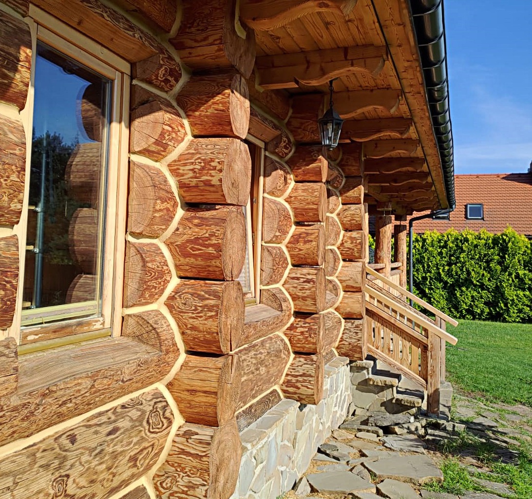 Ściana zewnętrzna domu z bali, widoczne okno z drewnianą ramą, kamienna podmurówka i wejście ze schodami, oświetlone słońcem.