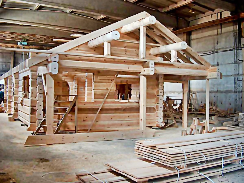 Surowa konstrukcja drewniana domu z bali w trakcie montażu w hali produkcyjnej, widoczne elementy dachu i ściany z wyciętymi otworami okiennymi, ułożone deski w dolnym rogu.