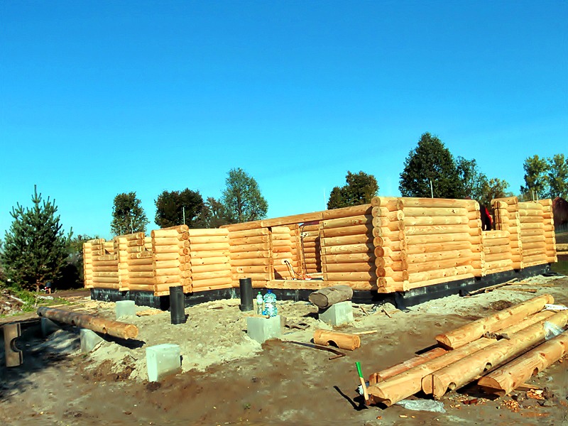 Wznoszona konstrukcja z bali na fundamencie, widoczne elementy izolacji i przygotowane bale na pierwszym planie, błękitne niebo w tle.