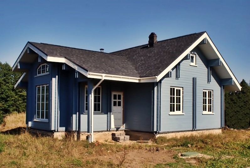 Niebieski dom szkieletowy z ciemnym dachem z gontu bitumicznego, białymi ramami okiennymi i drzwiami, widok z zewnątrz w słoneczny dzień, z widocznym kominem i elementami wykończeniowymi.