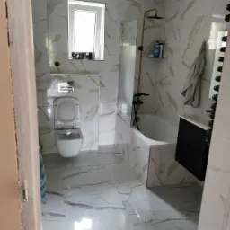 Wykończona łazienka z białymi płytkami imitującymi marmur z złotymi żyłami, z wiszącą toaletą, kabiną prysznicową typu walk-in z czarną armaturą i czarną szafką pod umywalką.