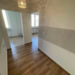 Korytarz po remoncie z brązowymi panelami imitującymi drewno, widok na pokój z oknem i grzejnikiem, na suficie lampa z ażurowym wzorem rzucająca cień na ścianę.