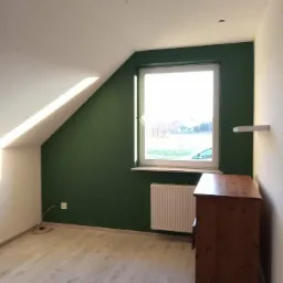 Pokój na poddaszu po remoncie: ściana pomalowana na ciemnozielono, drewniana komoda, jasne panele podłogowe, biały grzejnik pod oknem, skos dachu.