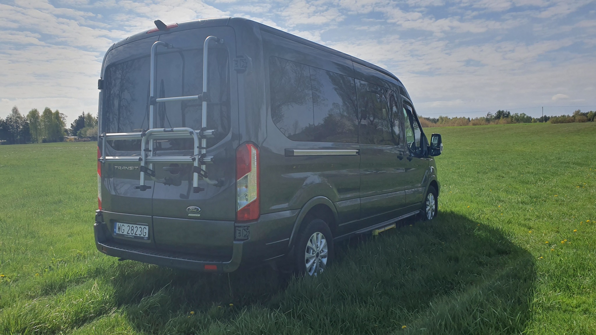Szary Ford Transit z bagażnikiem rowerowym z tyłu, zaparkowany na zielonej łące w słoneczny dzień.