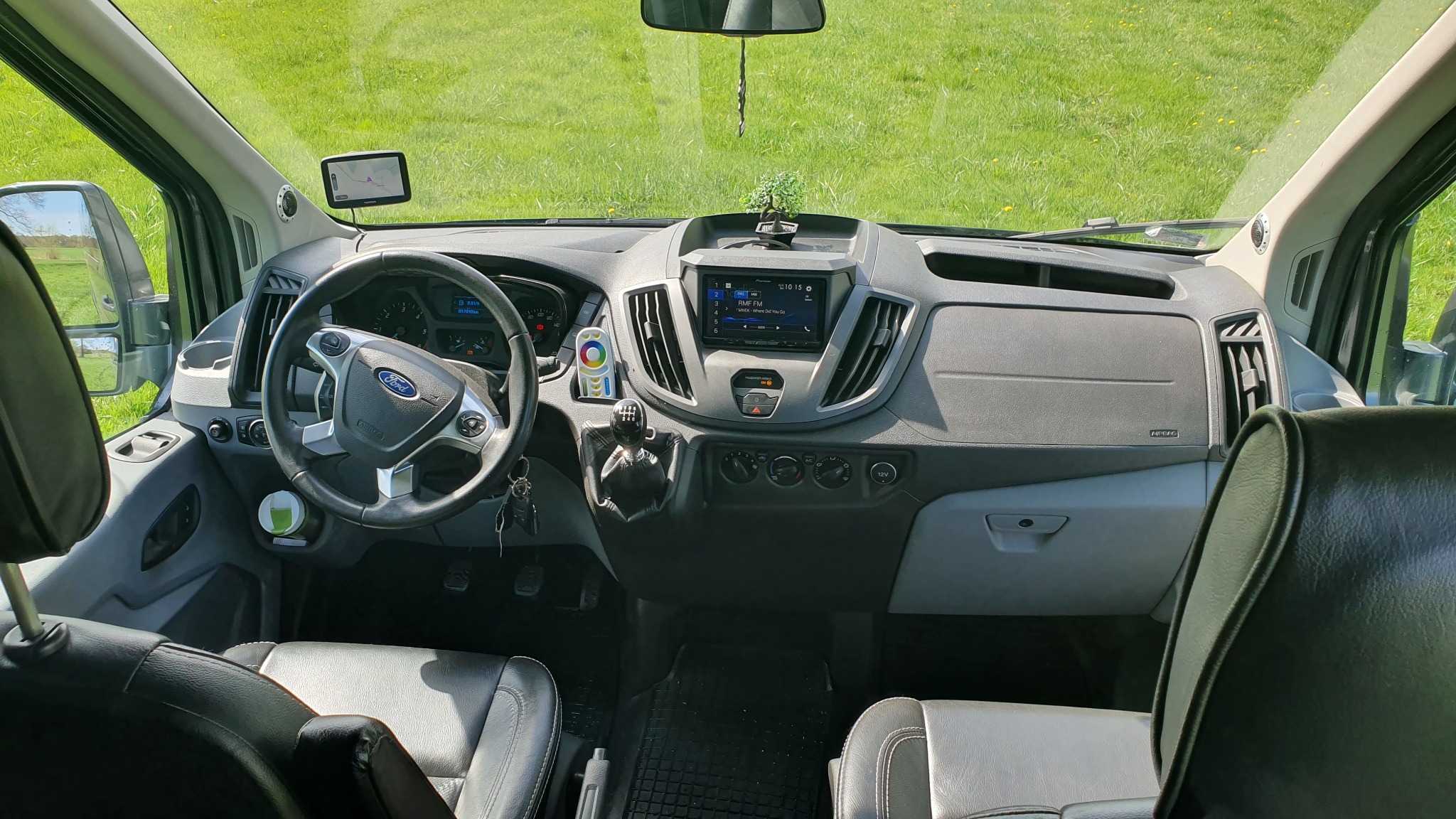 Wnętrze busa Ford Transit z widokiem na deskę rozdzielczą, kierownicę, siedzenia i zielone pole za szybą.