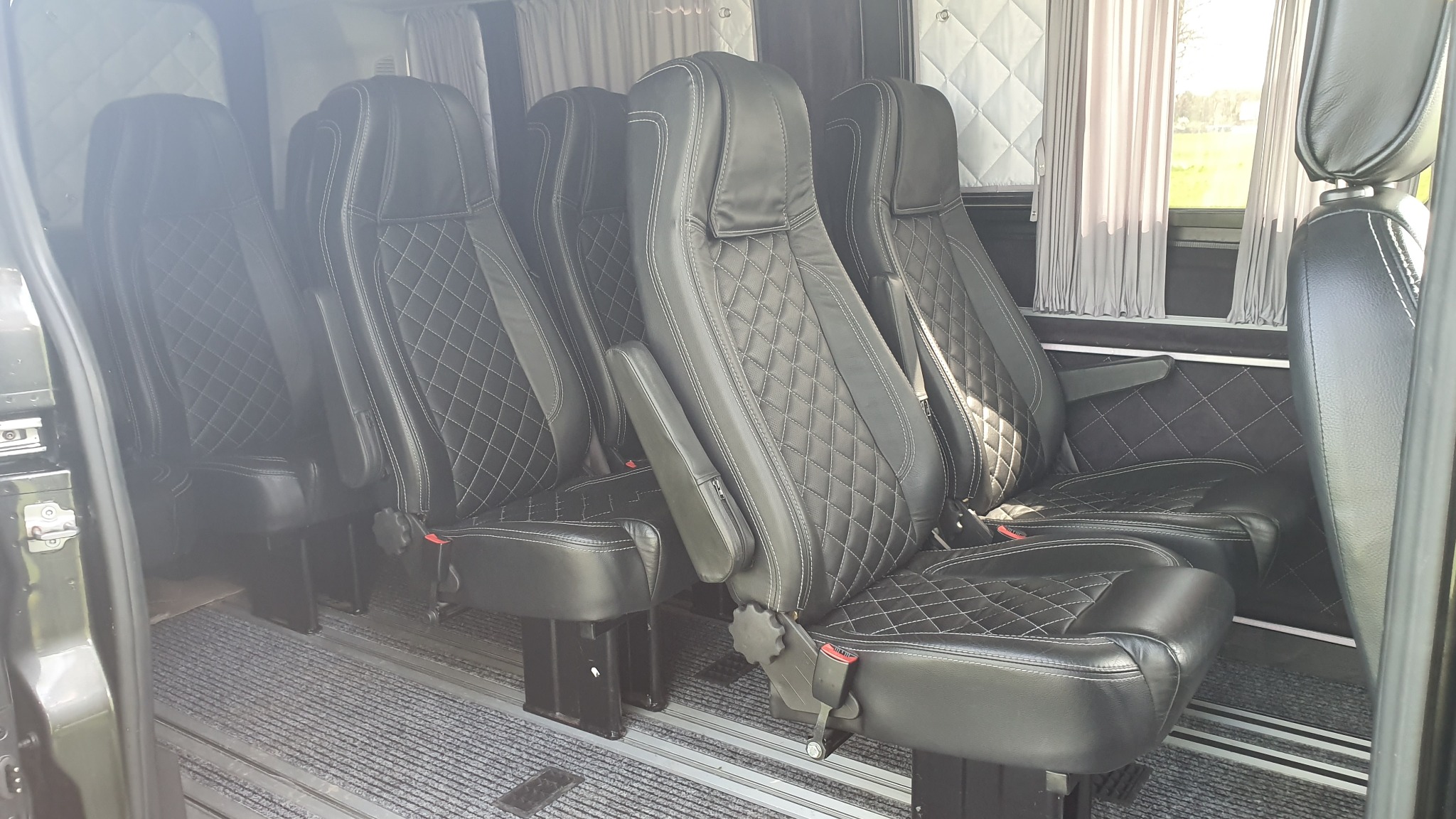 Wnętrze luksusowego busa z czarnymi, skórzanymi fotelami z białymi przeszyciami w romby, szare zasłonki w oknach i dywanik w paski.