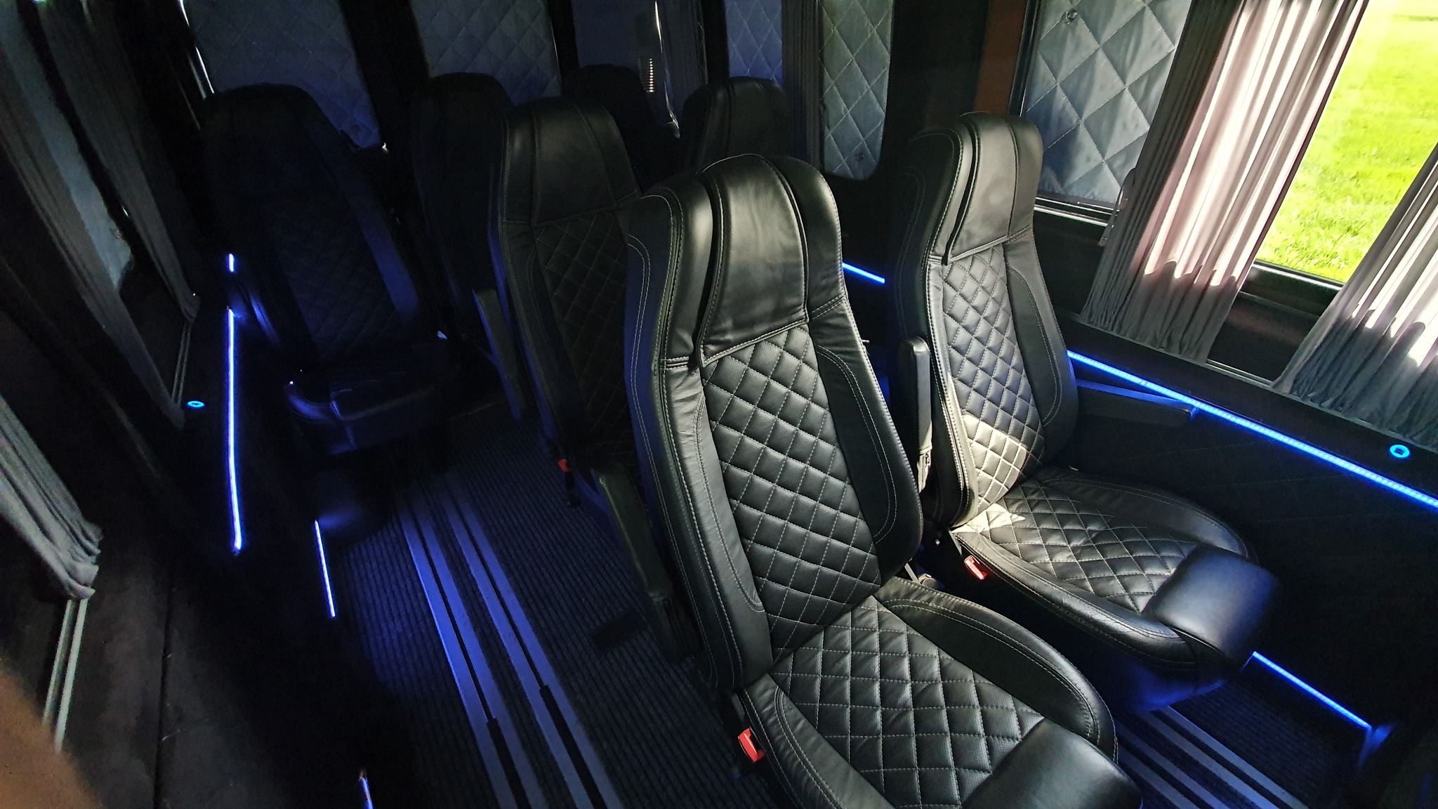 Wnętrze luksusowego busa z czarnymi skórzanymi fotelami w romby i niebieskim oświetleniem LED wzdłuż podłogi i okien.