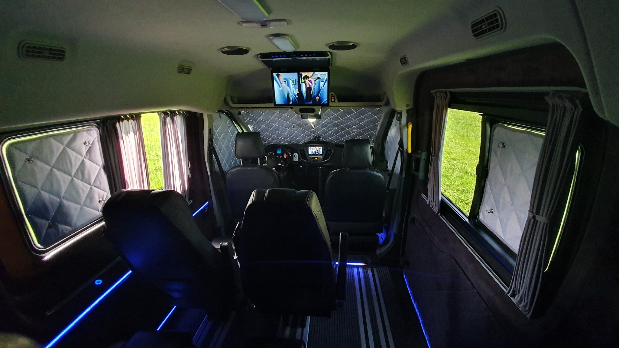 Wnętrze luksusowego busa z czarnymi skórzanymi fotelami, podświetleniem LED, zasłonami w oknach i telewizorem zamontowanym pod sufitem.