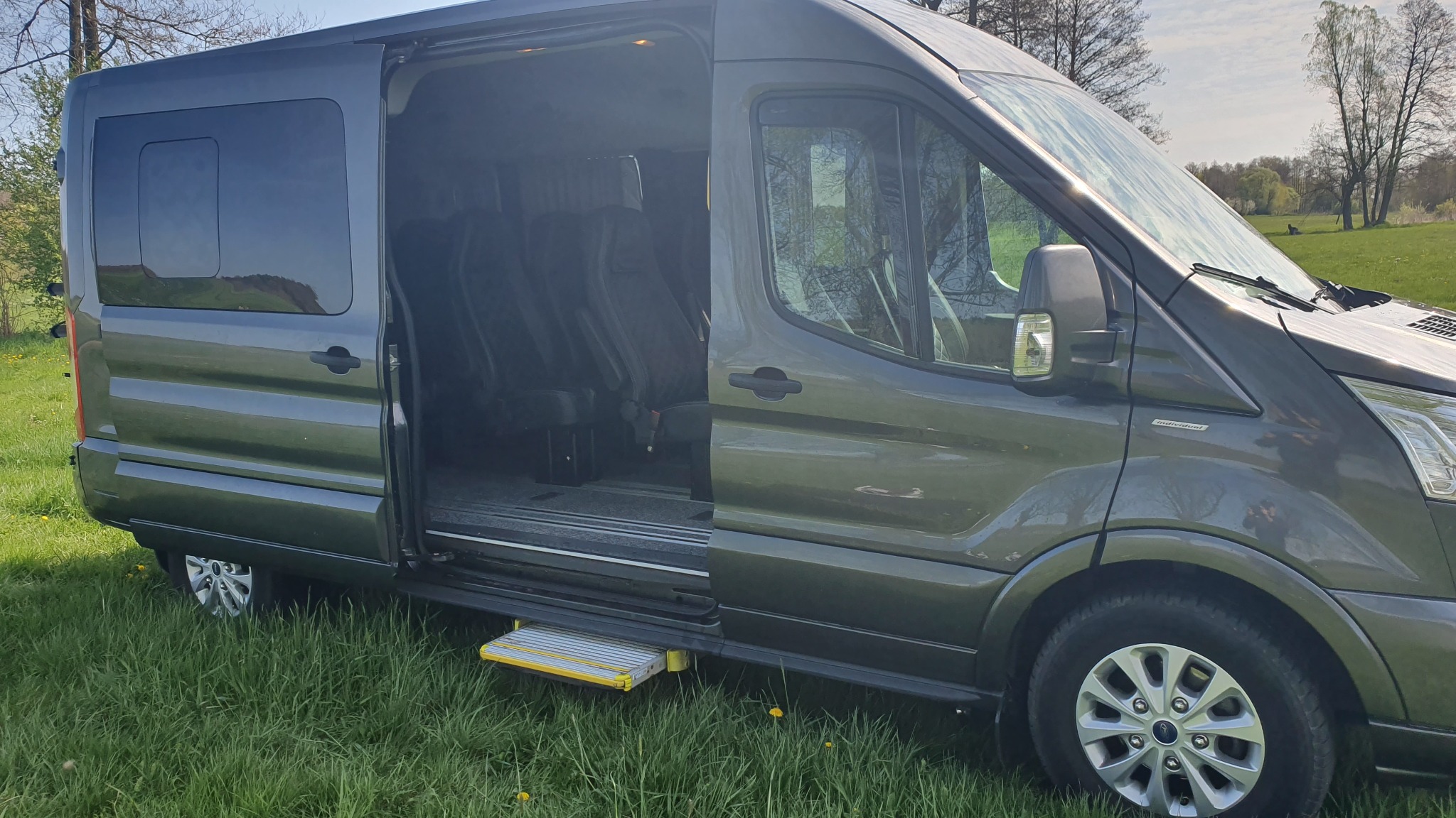 Szary van z otwartymi przesuwnymi drzwiami, widoczne wnętrze z rzędami foteli, stojący na trawiastym terenie.