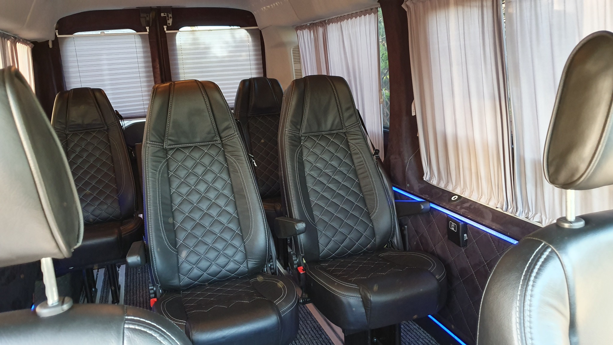 Wnętrze luksusowego busa z czarnymi skórzanymi fotelami z pikowaniem w romby, regulowane zagłówki, zasłony w oknach i nastrojowe niebieskie oświetlenie LED.