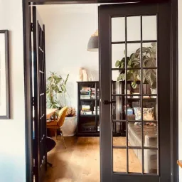 Czarne, przeszklone drzwi dwuskrzydłowe z widokiem na jasne wnętrze z drewnianą podłogą, regałem z książkami i roślinami doniczkowymi.