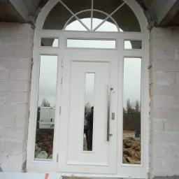 Białe drzwi wejściowe z półokrągłym nadświetlem i bocznymi szybami, zamontowane w surowym murze z betonu komórkowego, z widocznym odbiciem osób i otoczenia w szybach.