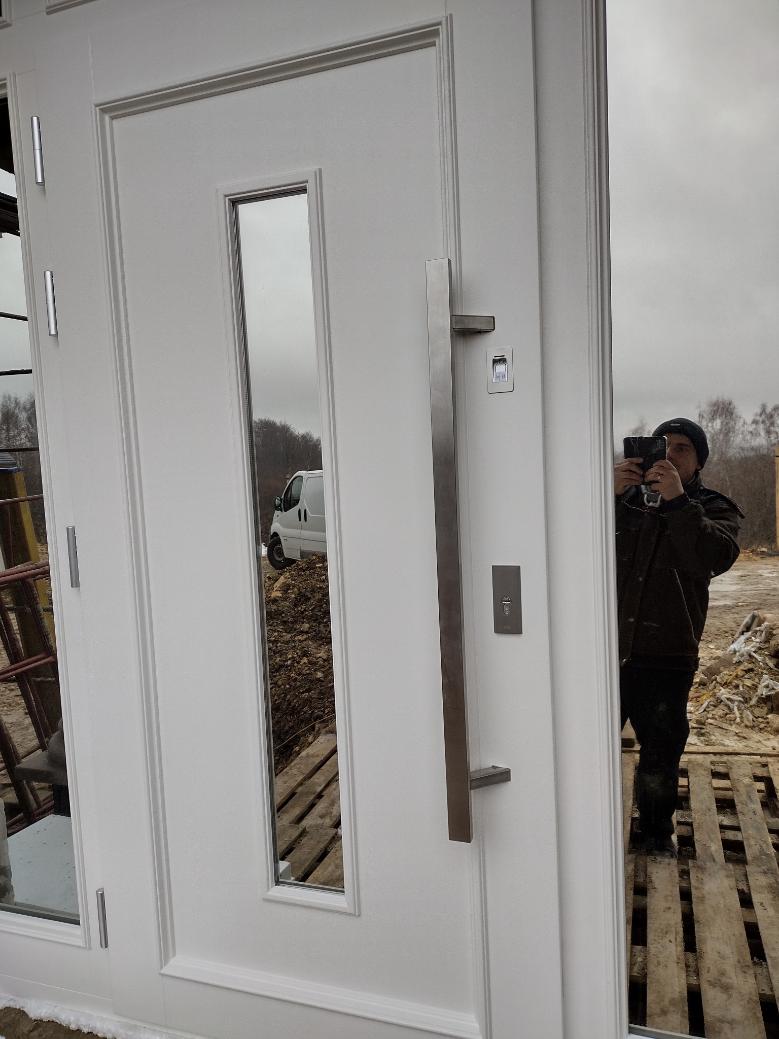Białe drzwi wejściowe z długim, pionowym uchwytem ze stali nierdzewnej i prostokątnym przeszkleniem, w którym odbija się biały samochód dostawczy oraz osoba robiąca zdjęcie telefonem.