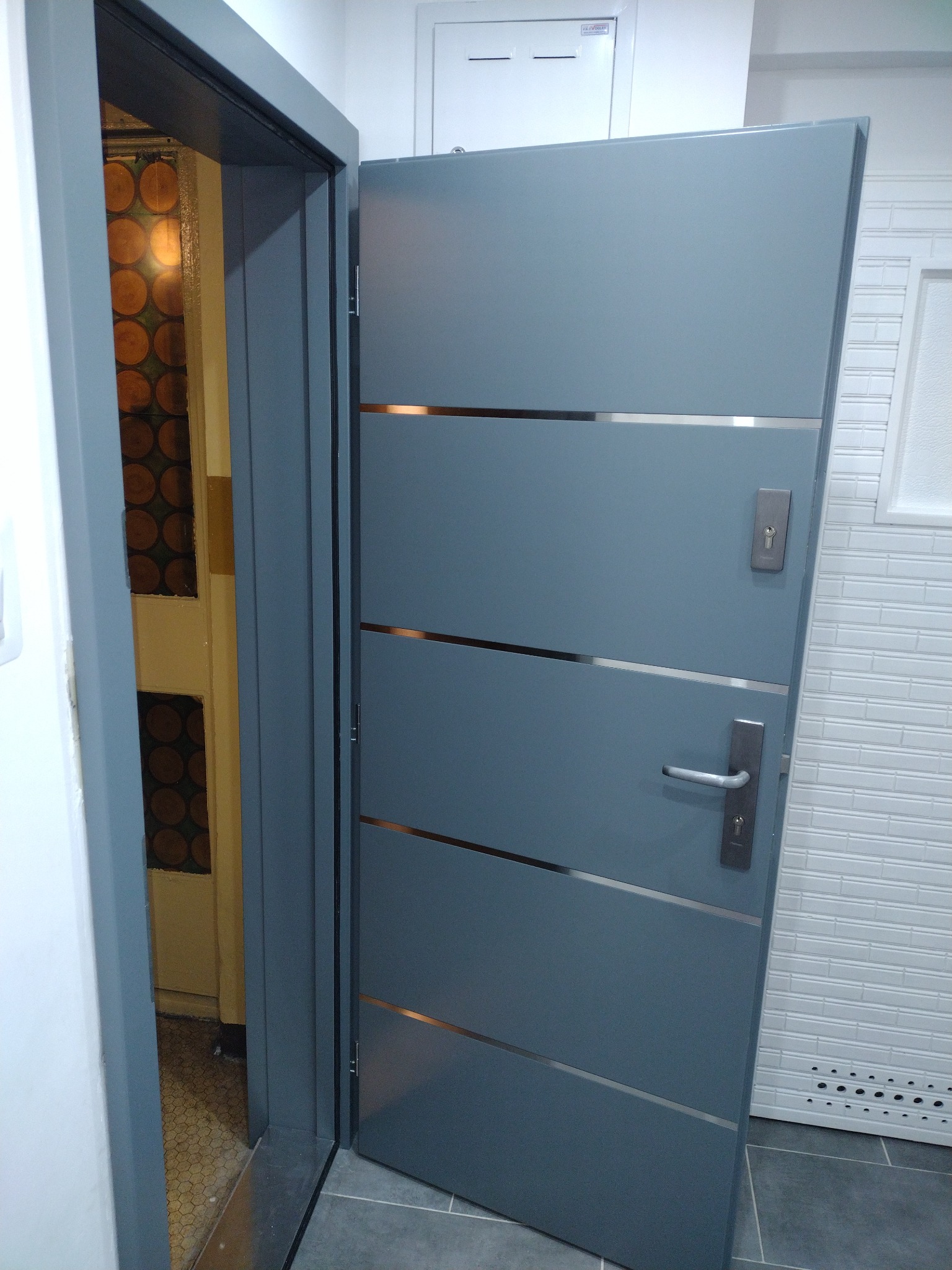 Otwarta, nowoczesna, szara drzwi wejściowe z poziomymi, metalowymi wstawkami, widoczny zamek i klamka, obok biała ściana z imitacją cegiełek i roleta.