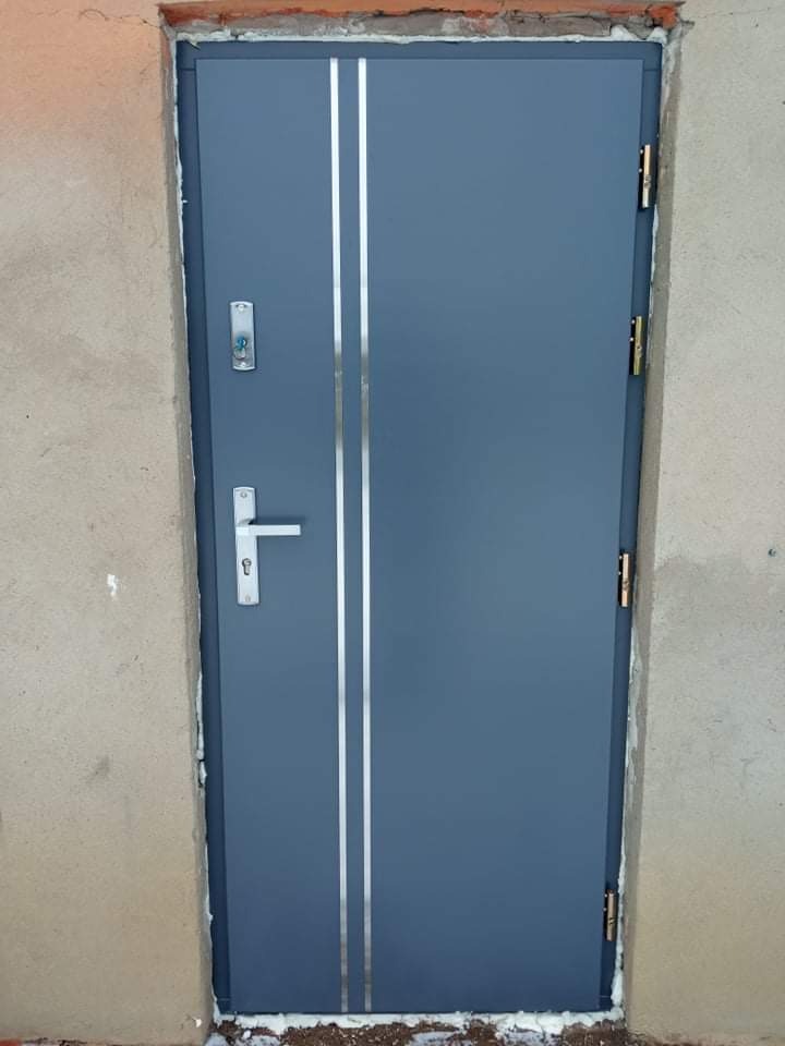 Szare, stalowe drzwi wejściowe z dwoma pionowymi, srebrnymi paskami, zamontowane w surowej, otynkowanej ścianie z widoczną pianką montażową.