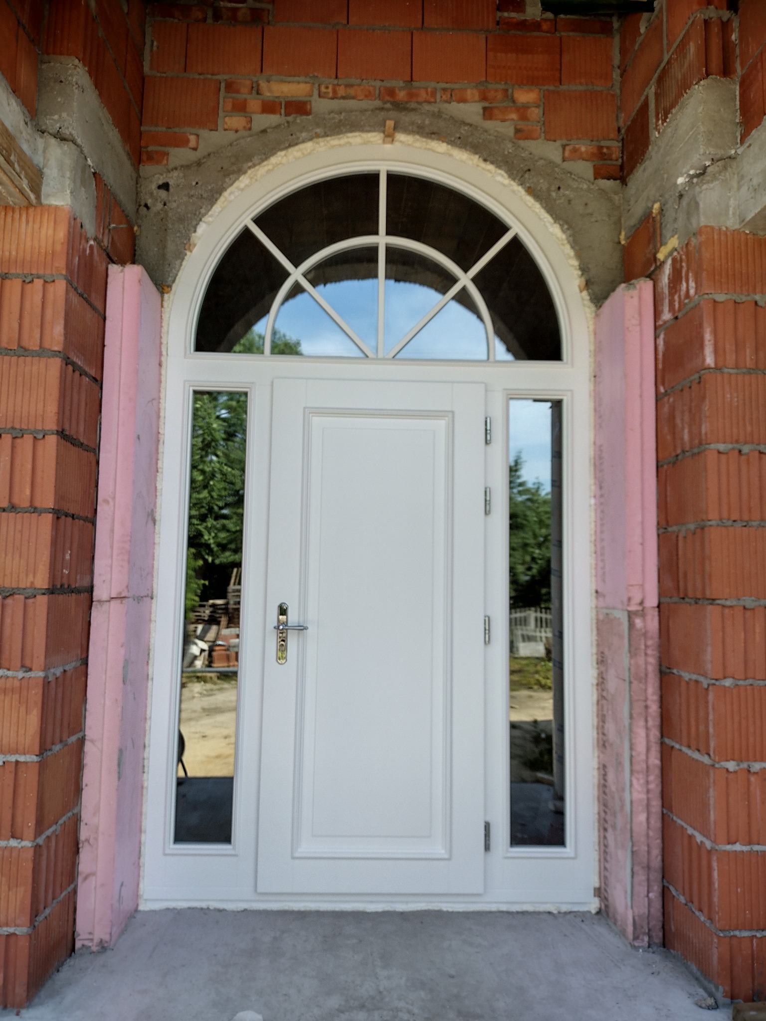 Białe drzwi wejściowe z półokrągłym nadświetlem, osadzone w surowym murze z czerwonej cegły, widoczne ocieplenie styropianem po bokach.