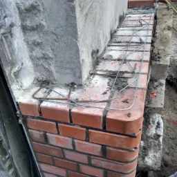 Ceglana obudowa zbrojona drutem na fundamencie, widoczne fragmenty betonu i ziemi.