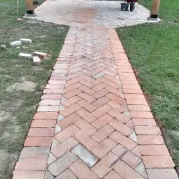 Ułożona z cegły ścieżka w jodełkę prowadząca do drewnianej altany, taczka z narzędziami w tle, trawnik po bokach.