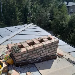 Wznoszenie komina z cegły na dachu pokrytym blachą, widoczne narzędzia i materiały budowlane, w tle zieleń drzew.