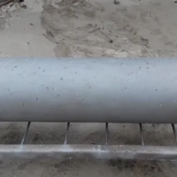 Cylindryczny element po obróbce, leżący na metalowej konstrukcji, z widoczną oponą w tle.