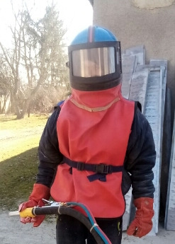 Osoba w kombinezonie ochronnym i masce do piaskowania, trzymająca pistolet do piaskowania, z widocznymi przewodami, na tle drzew i elementów konstrukcyjnych.