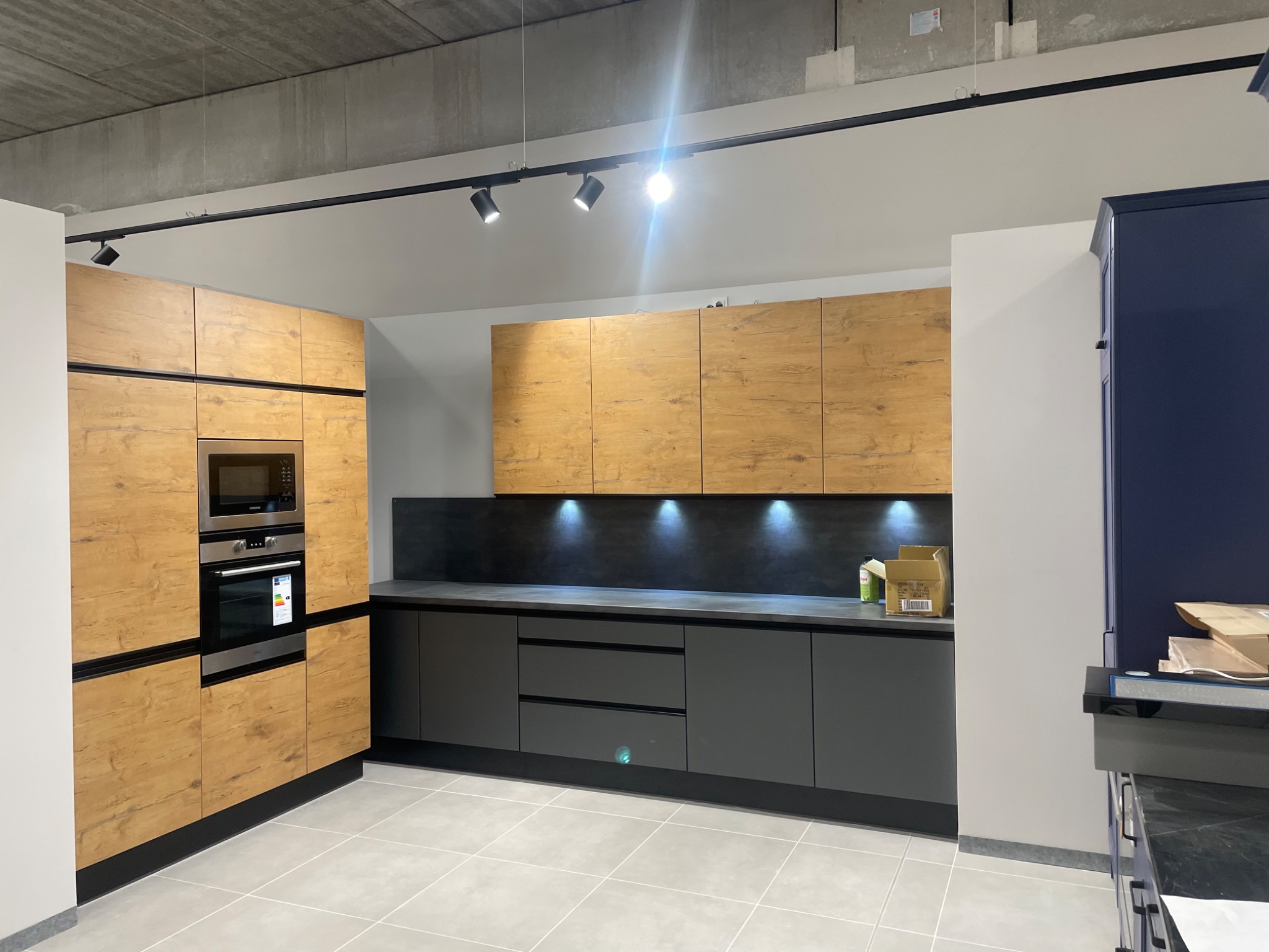 Nowoczesna kuchnia z drewnianymi frontami szafek górnych i szarymi dolnymi, oświetlona punktowymi lampami na szynie, z wbudowanym piekarnikiem i mikrofalówką, na tle surowego, betonowego sufitu.