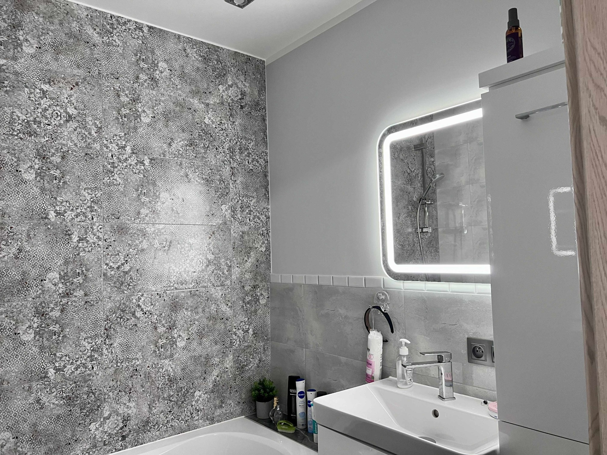 Nowoczesna łazienka w szarościach z wzorzystą płytką na ścianie, podświetlanym lustrem i białą umywalką. Widoczne detale wyposażenia i oświetlenie.