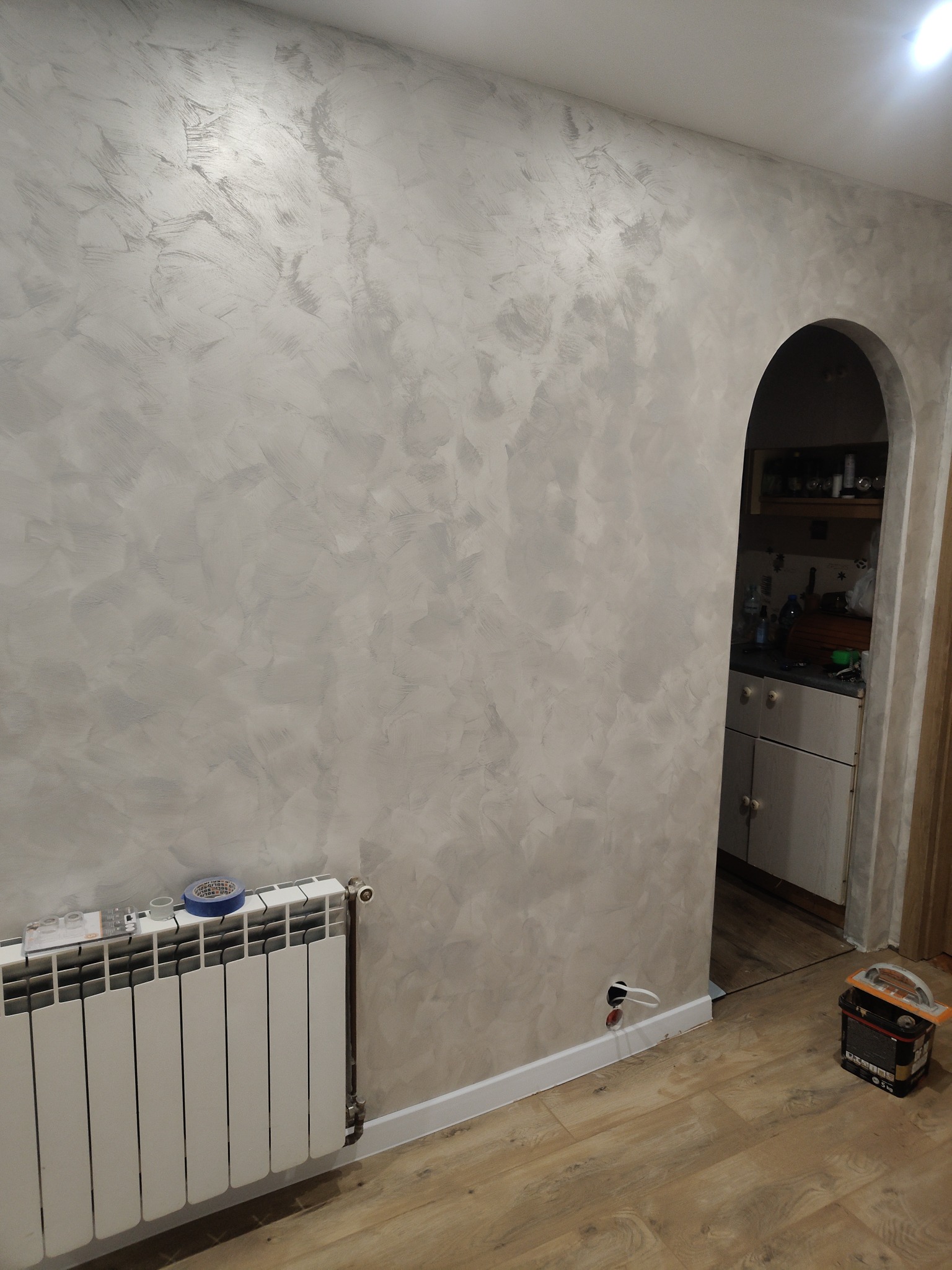 Ściana z dekoracyjną szarą fakturą, widoczny grzejnik i łukowate przejście do kuchni. Na podłodze leżą narzędzia malarskie.