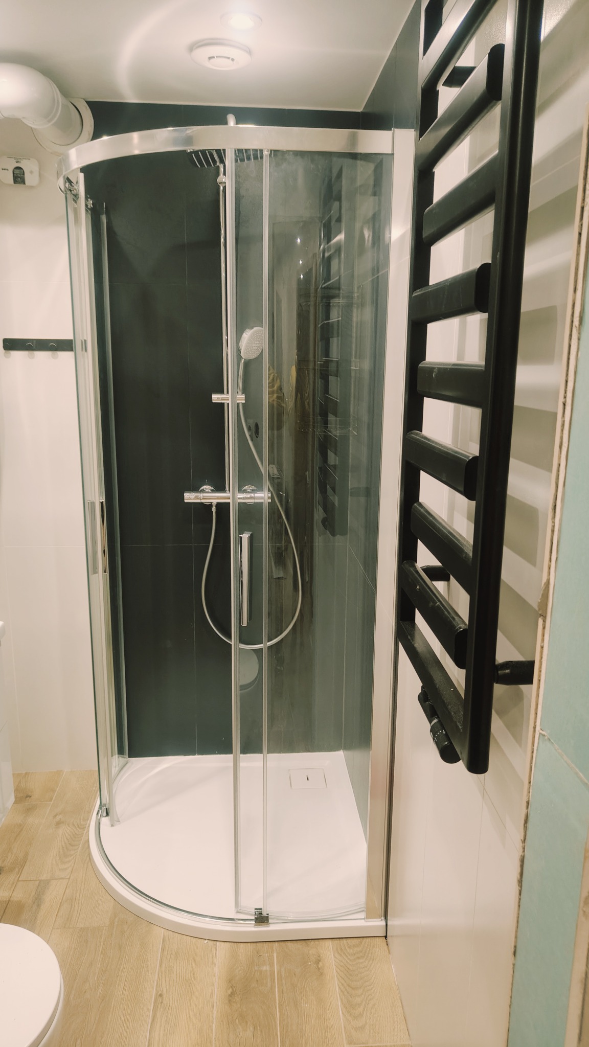 Nowoczesna kabina prysznicowa z czarną ścianą i grzejnikiem drabinkowym, widok z boku, na podłodze panele imitujące drewno, białe ściany, oświetlenie sufitowe.