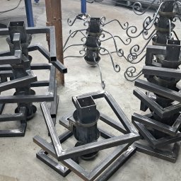 ArtisOmnia - Nieskończone metalowe konstrukcje spawane z profili kwadratowych, tworzące geometryczne formy przypominające podstawy lamp lub kwietników, widoczne w warsztacie z narzędziami i skrzynią...