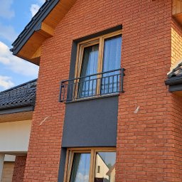 ArtisOmnia - Nowoczesny dom z cegły klinkierowej z czarną balustradą balkonową i oknami w drewnianych ramach, widok z dołu na tle błękitnego nieba z chmurami.