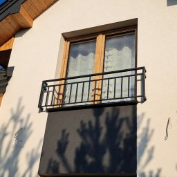 ArtisOmnia - Czarna, prosta balustrada balkonowa z metalu, zamontowana przy oknie na elewacji budynku o jasnej fasadzie i drewnianych elementach dachu.