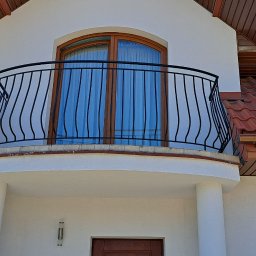 ArtisOmnia - Czarna, ozdobna balustrada balkonowa z pionowymi prętami na białym budynku z brązowym dachem i oknami dachowymi.