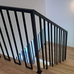 ArtisOmnia - Nowoczesna, czarna balustrada schodowa z pionowymi szczeblami w minimalistycznym wnętrzu z drewnianymi schodami i białymi ścianami, oświetlona punktowym światłem.